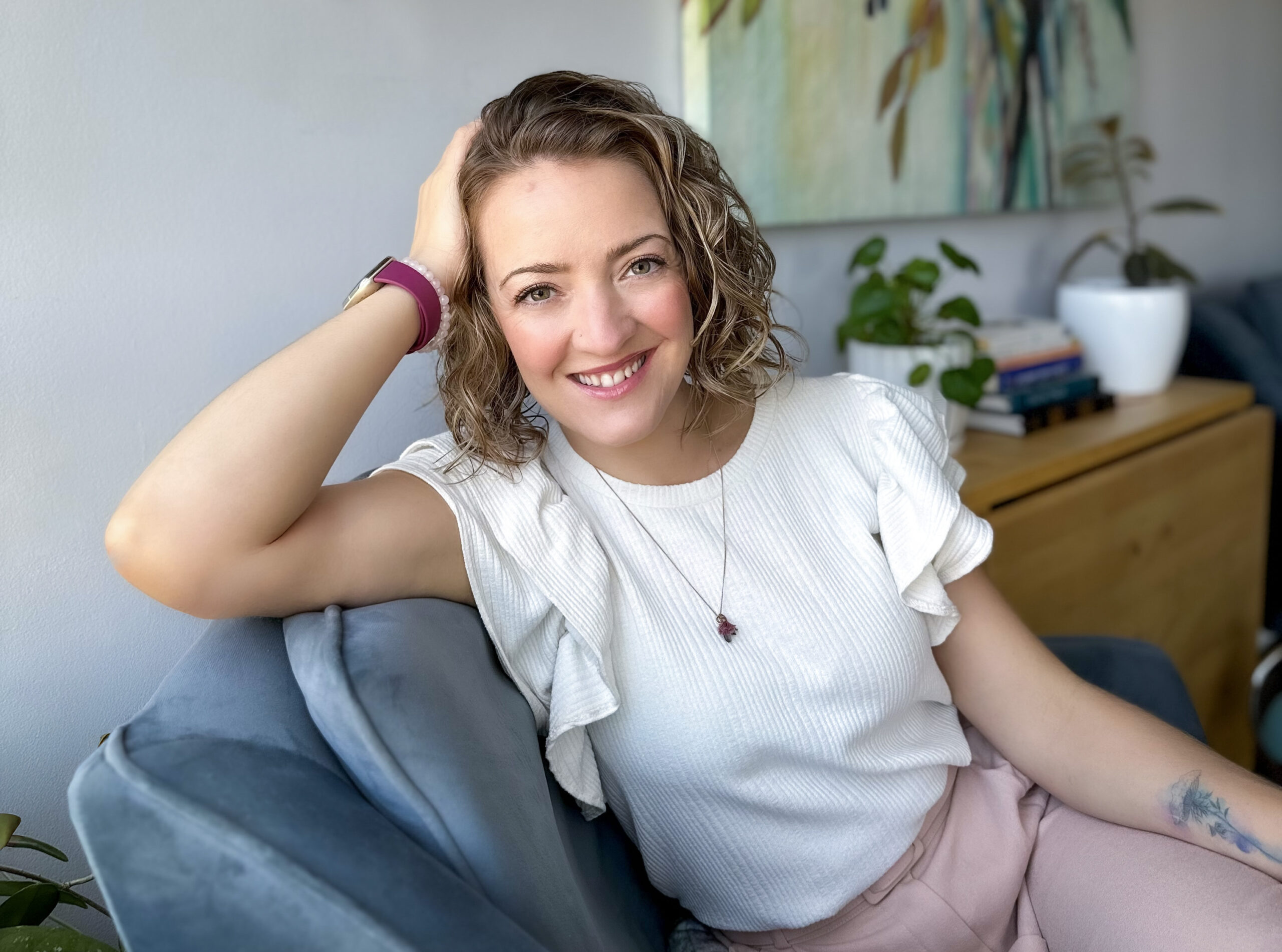 A white female mental health professional smiles while sitting in a chair and leaning toward you with her head leaning against her right hand.